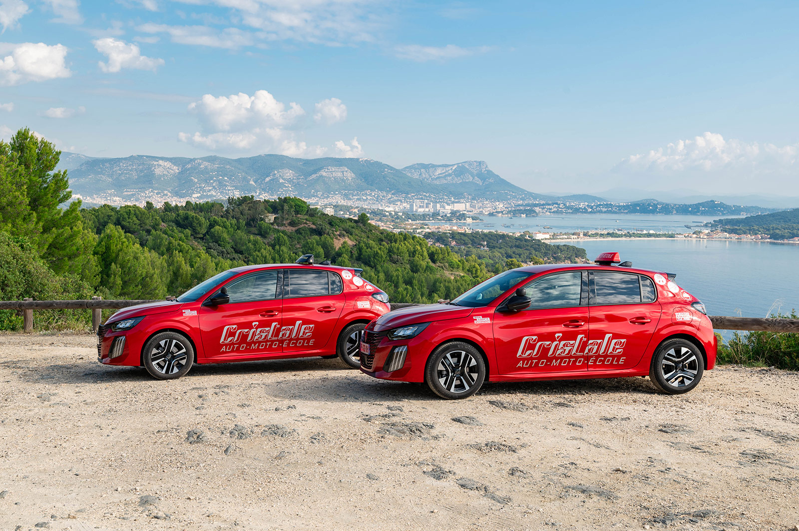 2 automobiles rouges de l'auto-moto école Cristale à la Seyne-sur-Mer sur un parking surplombant la rade de Toulon