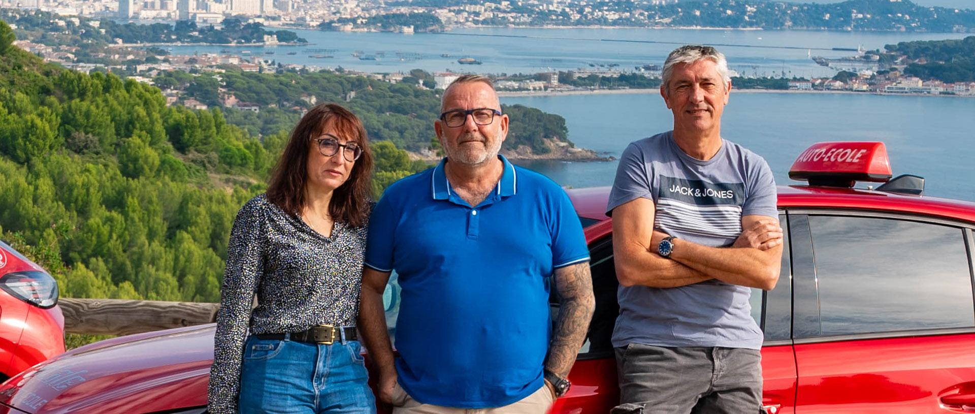 Les 3 moniteurs de l'auto-moto école Cristale à la Seyne-sur-Mer devant 2 automobiles rouges sur un parking surplombant la rade de Toulon